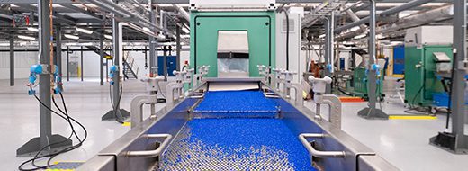 A factory interior with a conveyor belt transporting blue plastic pellets toward a large green industrial machine, surrounded by pipes and equipment.