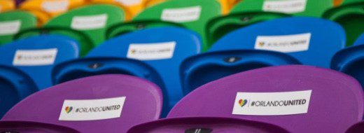 Rows of empty stadium seats in rainbow colors, each with a sticker displaying a heart and the hashtag #ORLANDOUNITED.