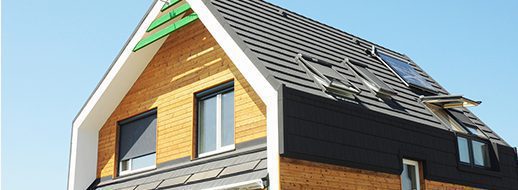 Modern house with a steep black roof, wooden siding, large windows, and skylights, photographed under a clear blue sky.