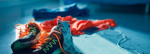 A pair of running shoes with orange laces sits on the floor next to scattered clothes and a tangled charging cable in a sunlit room.