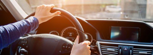 A person drives a car, holding the steering wheel with one hand. The dashboard and sunlight through the windshield are visible.