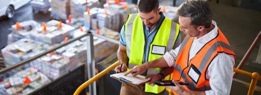 Two workers wearing safety vests review documents on a clipboard at a warehouse, with stacked goods and boxes visible in the background.