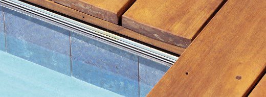 Close-up of a wooden deck next to the edge of a tiled swimming pool, showing the water and pool wall tiles beneath the deck.