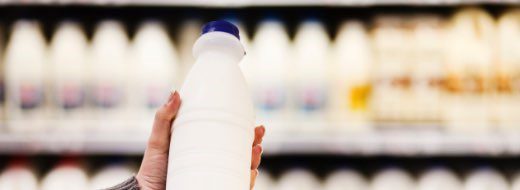 A hand is holding a white plastic milk bottle with a blue cap in front of a refrigerated display of similar bottles in a store.