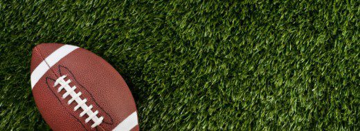 A brown American football with white laces and stripes lies on green artificial turf.