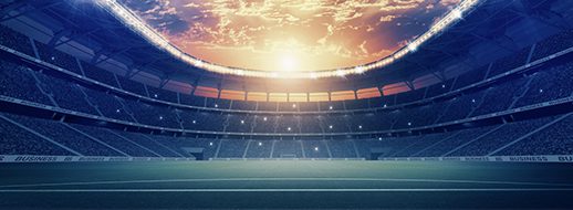 Large, empty sports stadium under a dramatic sky at sunset, with seats surrounding a well-lit field and bright lights illuminating the arena.