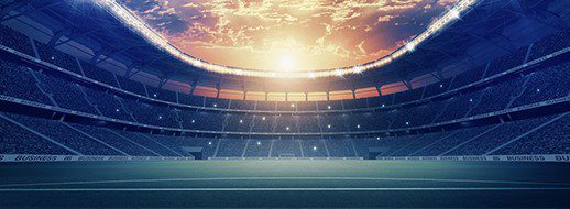 Empty sports stadium with a green field and rows of seats, illuminated under a dramatic sunset sky.