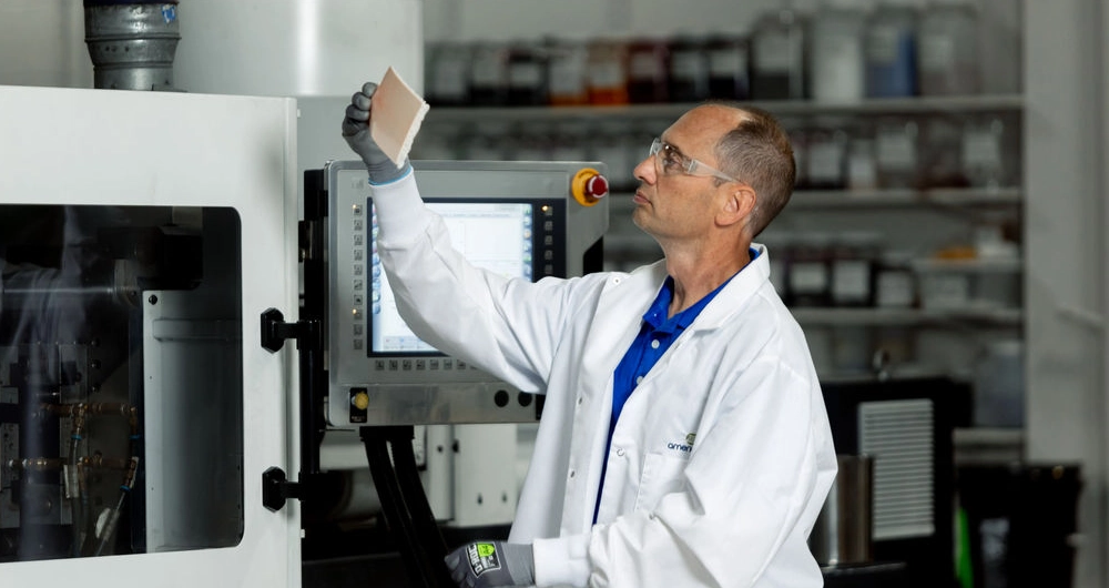 A man in a white lab coat and gloves examines a rectangular sample in front of industrial equipment inside a laboratory or manufacturing facility.