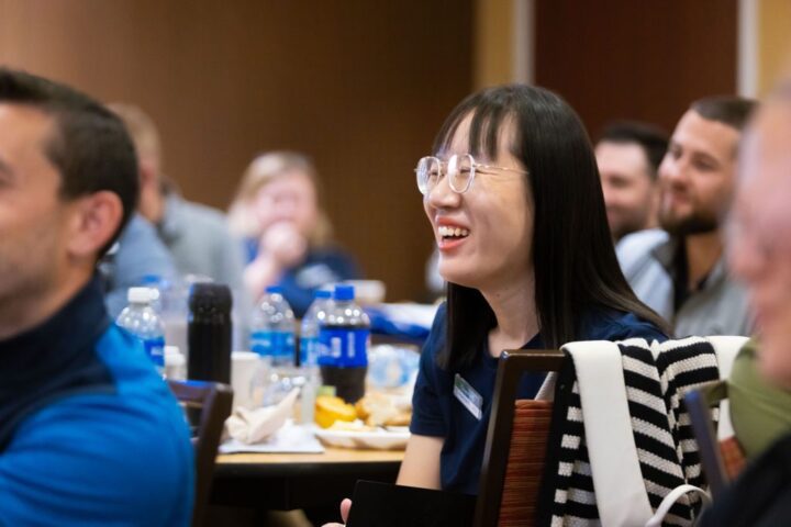 A woman with glasses smiles while seated at a table with water bottles and food, surrounded by other people in a conference or meeting setting.