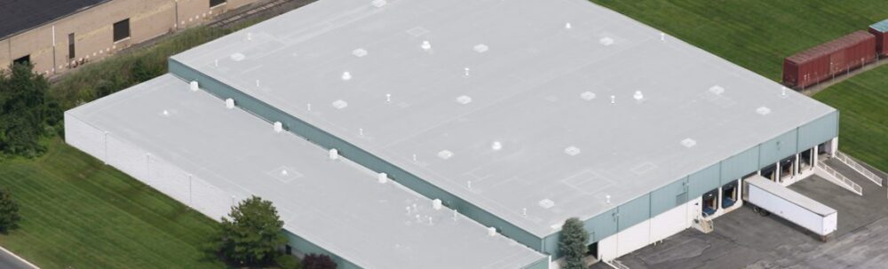 Aerial view of a large industrial warehouse with a flat white roof, multiple loading docks, and surrounding green grass.