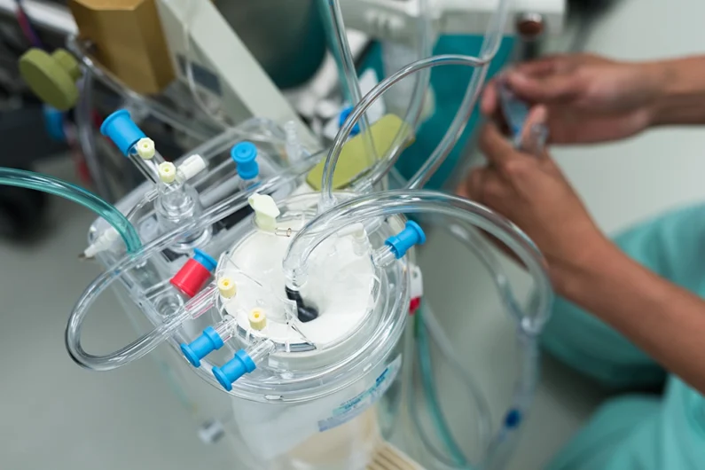 Close-up of a medical device with tubes and connectors, possibly an extracorporeal circuit, with a person's hands adjusting equipment in the background.