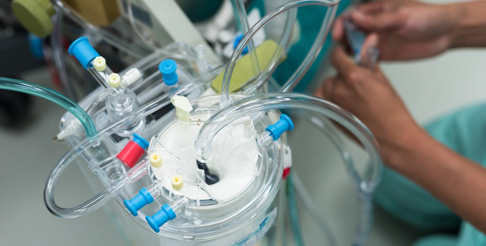 Close-up view of a heart-lung machine with transparent tubes, connectors, and medical components; a person adjusts equipment in the background.