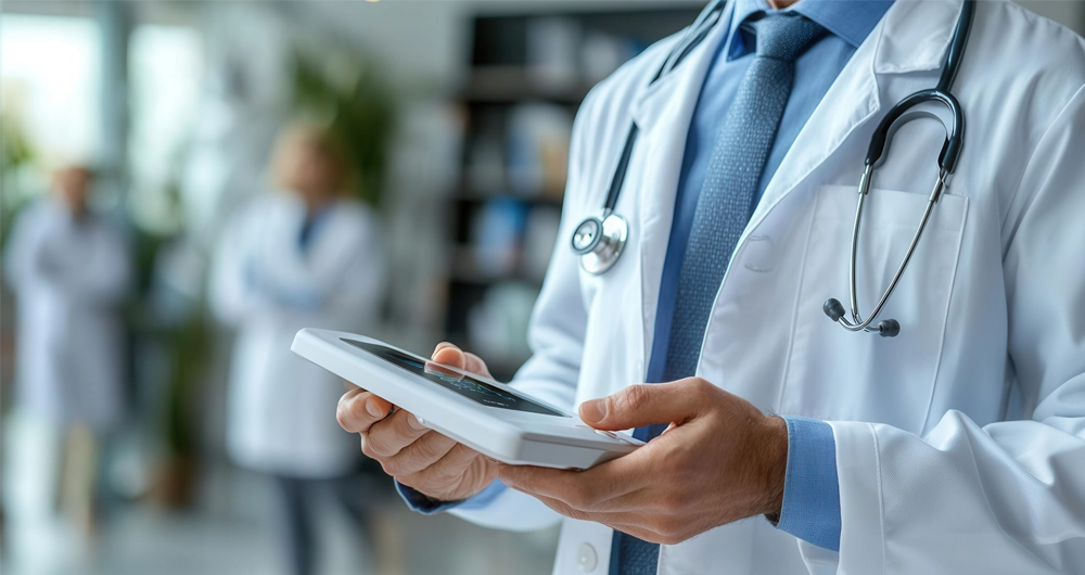 A doctor in a white coat with a stethoscope uses a tablet device in a medical setting, with two blurred figures in the background.