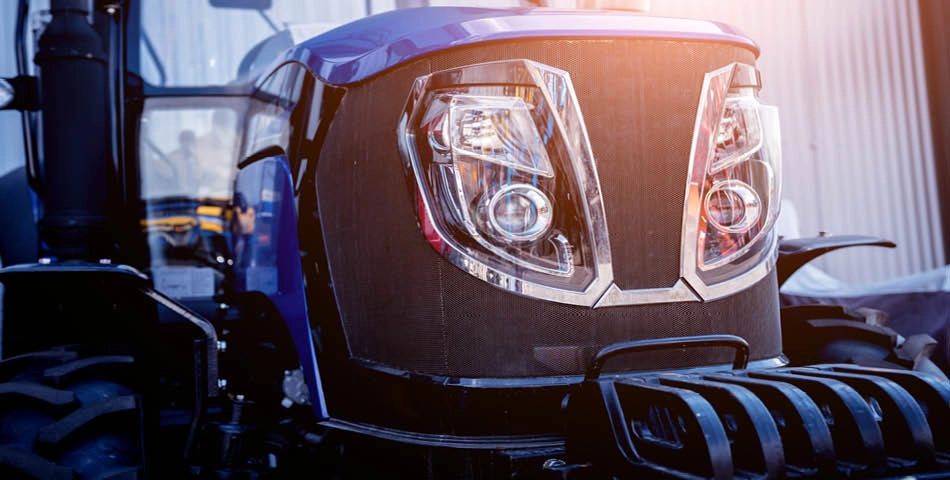 Close-up view of the front of a blue tractor, showing headlights, grille, and part of the large tires, with sunlight shining from the upper right.