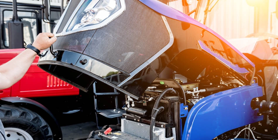 A person lifts the hood of a blue tractor, revealing its engine and internal components in an outdoor setting.