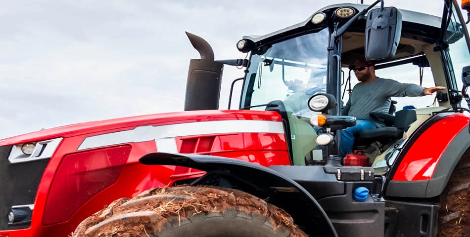 A person wearing sunglasses sits inside the cab of a modern red tractor with muddy tires, outdoors under a cloudy sky.