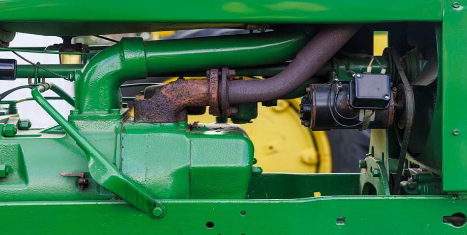 Close-up view of a green tractor engine showing metal pipes, a rusty exhaust component, and various mechanical parts.