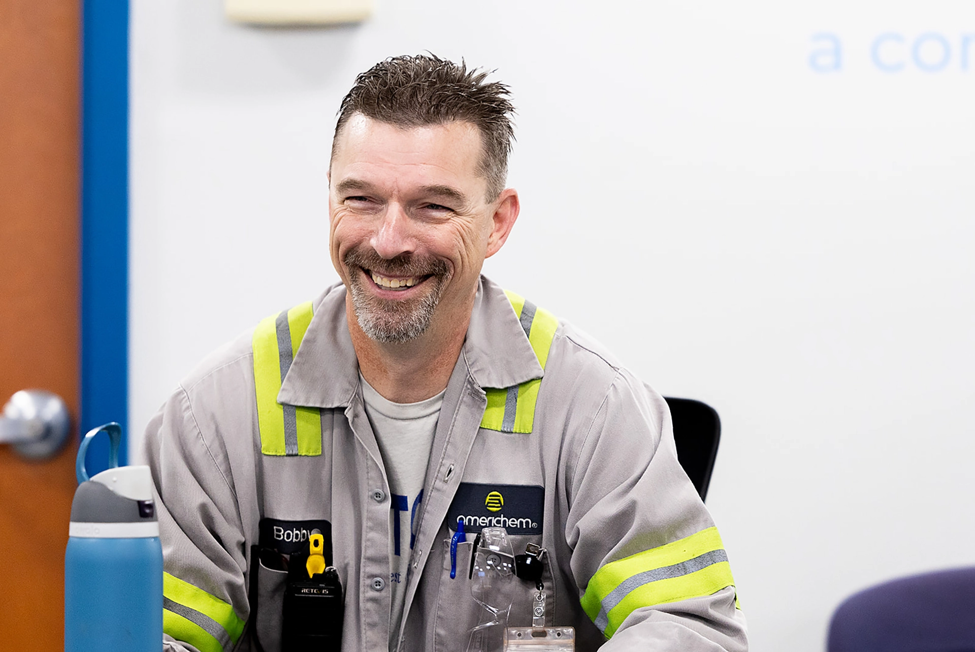 A man in a work uniform with reflective stripes sits at a table, smiling. He has short hair, a goatee, and a water bottle in front of him.