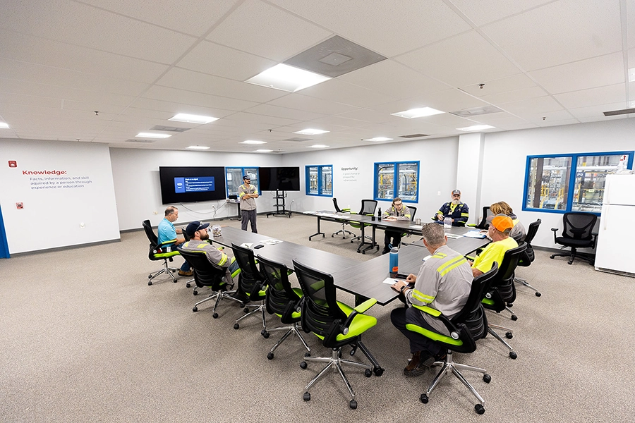 A group of workers in safety gear sit around tables in a conference room while a person gives a presentation in front of large monitors.