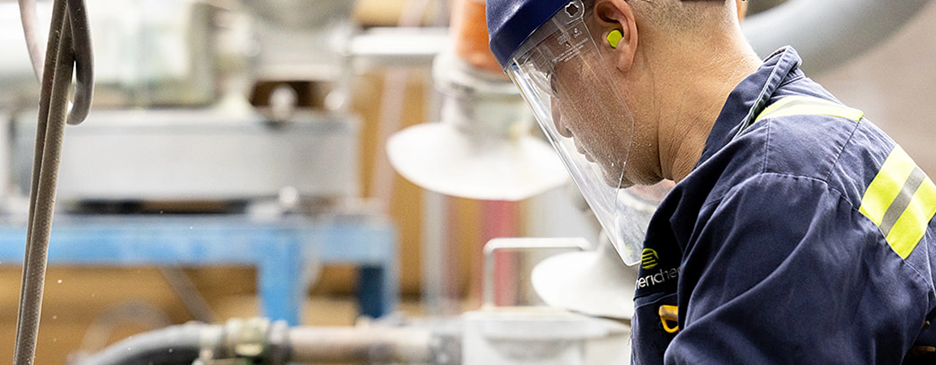 A worker wearing protective gear, including a face shield, earplugs, and a blue uniform, operates machinery in an industrial setting.