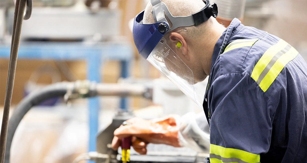 A worker wearing safety gear, including a face shield, earplugs, gloves, and a reflective jacket, operates machinery in an industrial setting.