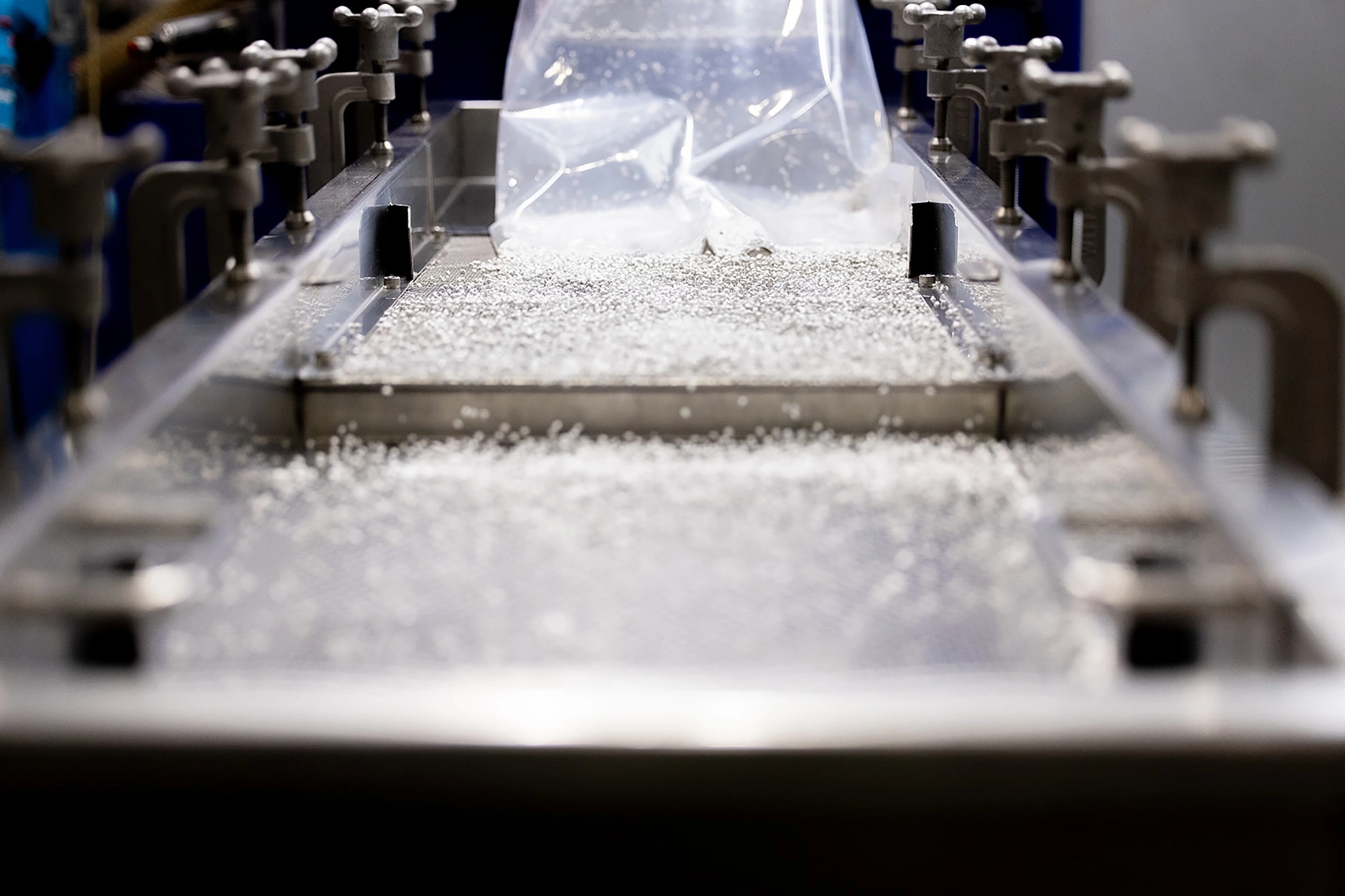 Clear plastic pellets are dispensed onto a metal surface in an industrial setting, with clamps lining both sides of the machine.