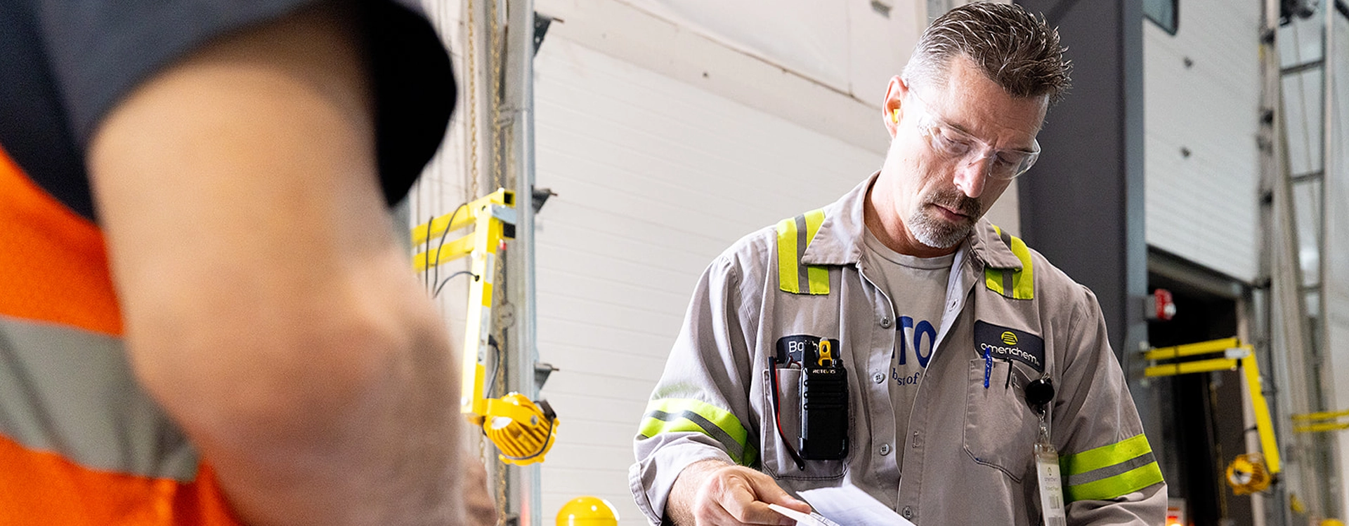 A man in safety glasses and a reflective work jacket reviews paperwork in an industrial or warehouse setting. Another person in an orange safety vest is partially visible in the foreground.