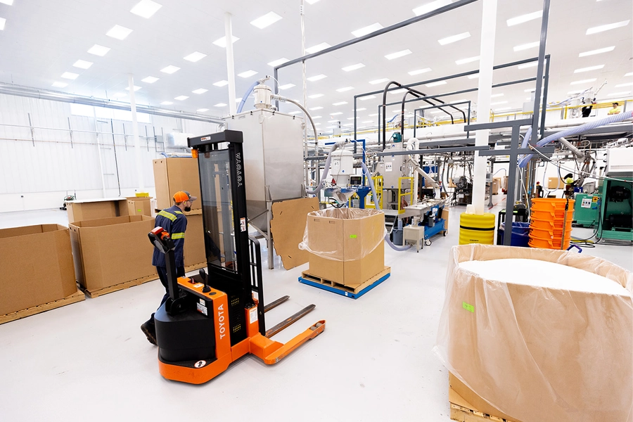 A worker operates an orange Toyota electric pallet jack in a clean, brightly lit industrial facility with large boxes and machinery.