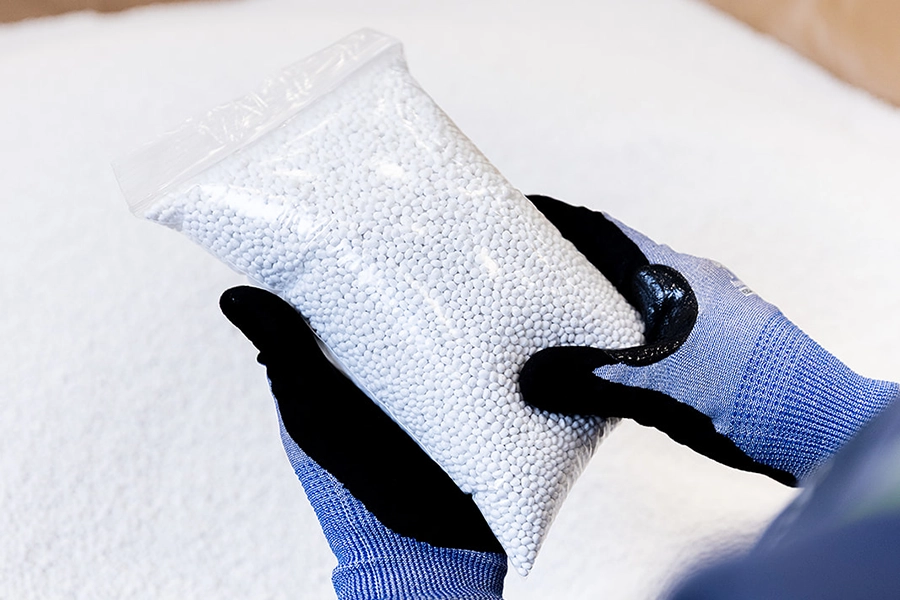 A person wearing blue gloves holds a clear plastic bag filled with small white plastic pellets.