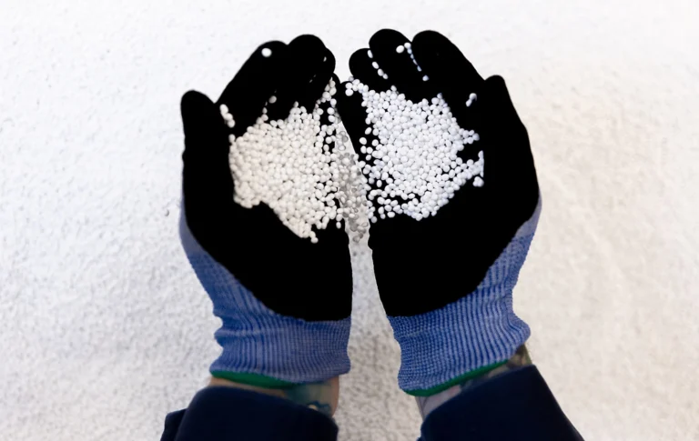 A person wearing blue and black gloves holds small white pellets in their cupped hands over a surface covered with similar pellets.