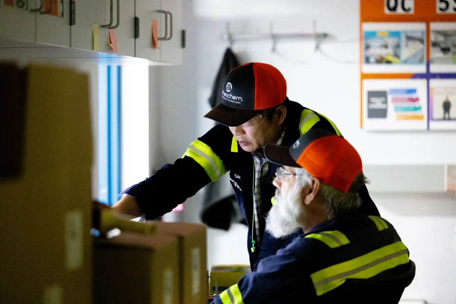 Two workers in uniforms and orange caps examine documents and boxes in an office, with calendars and notice boards visible on the wall in the background.