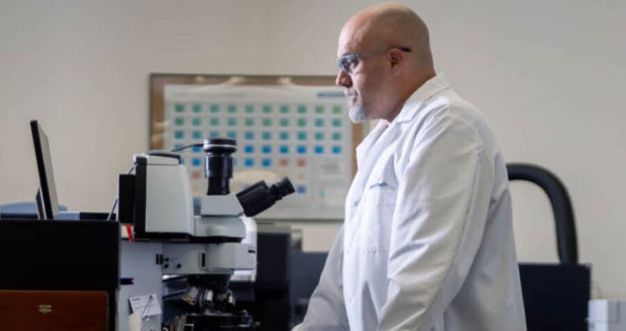 A person in a lab coat and safety glasses stands in a laboratory, working at a computer next to a microscope.