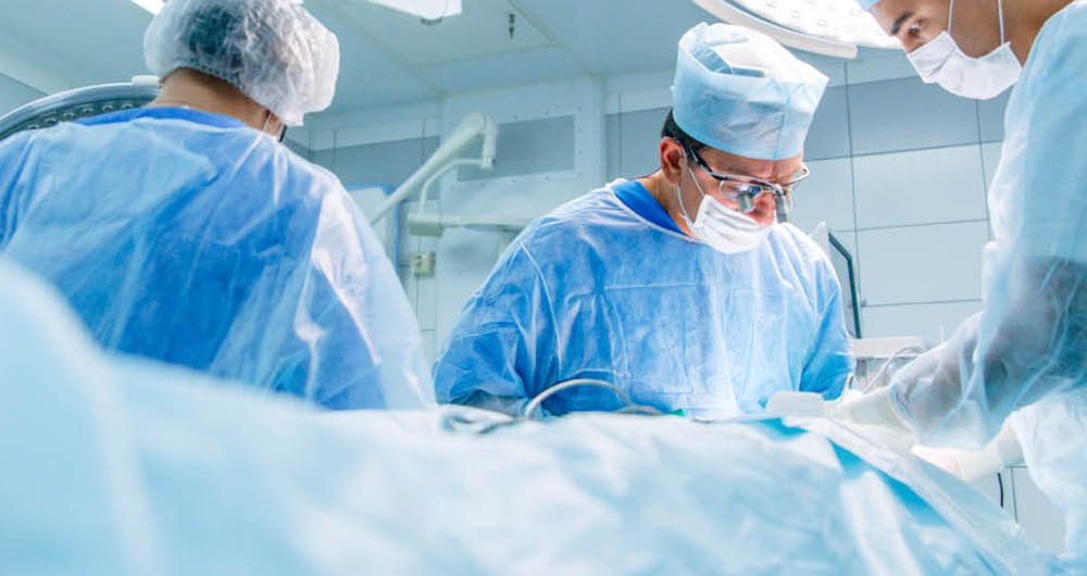 Three surgeons in scrubs and masks perform a medical procedure in a well-lit operating room.