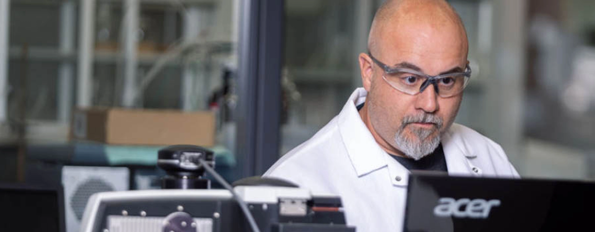 A person wearing safety glasses and a lab coat works at a computer in a laboratory setting.