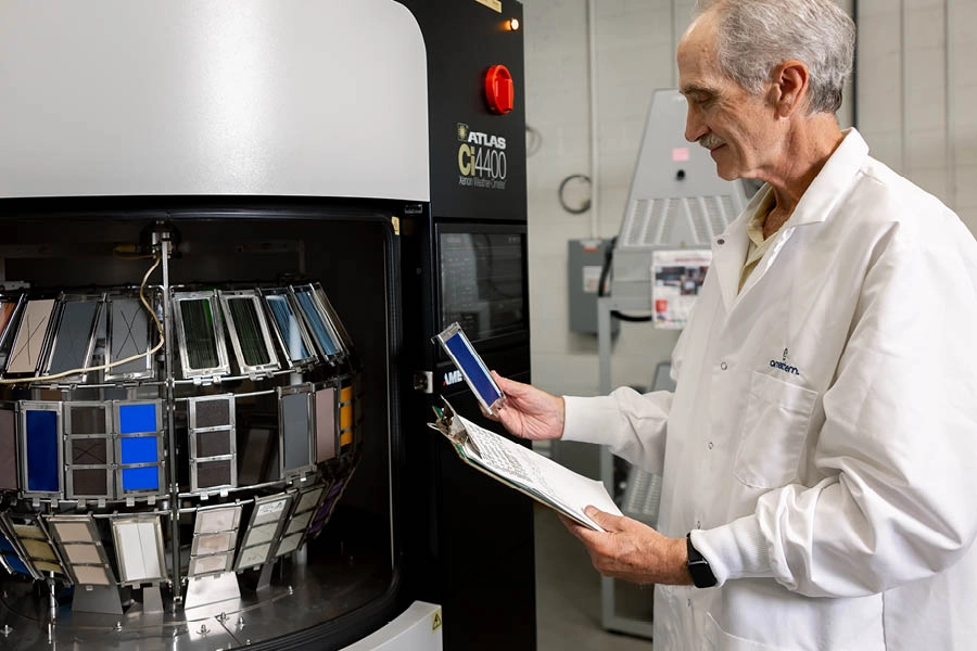 A scientist in a lab coat examines color test samples and records notes next to a large industrial testing machine.