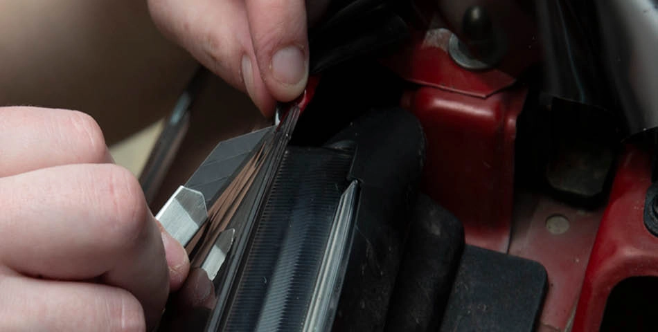 Close-up of hands using a utility knife to cut a sheet of window tint film being applied to a car window.
