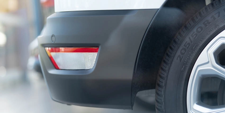 Close-up of a car’s rear bumper showing a red and white reflector, black plastic trim, tire, and part of the wheel with visible tire size markings.
