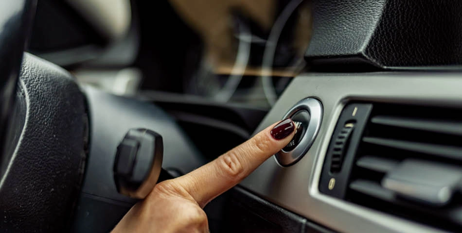 A person with dark red nail polish presses the start button on a car’s dashboard, next to the steering wheel and air vent.