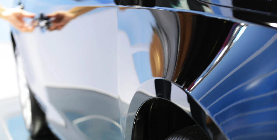 Close-up of a shiny black car door with a person’s hands reaching for the handle, reflecting surroundings on the vehicle's surface.