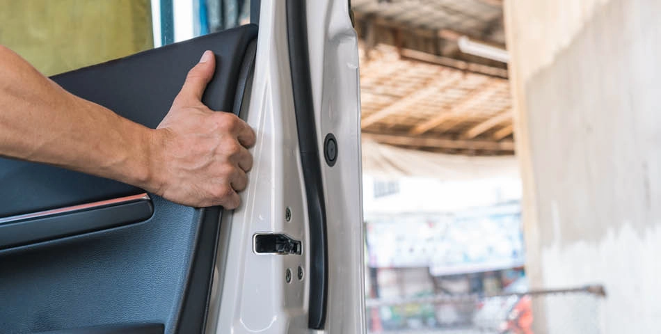 A person’s hand is opening the interior side of a car door, with part of an outdoor area visible in the background.