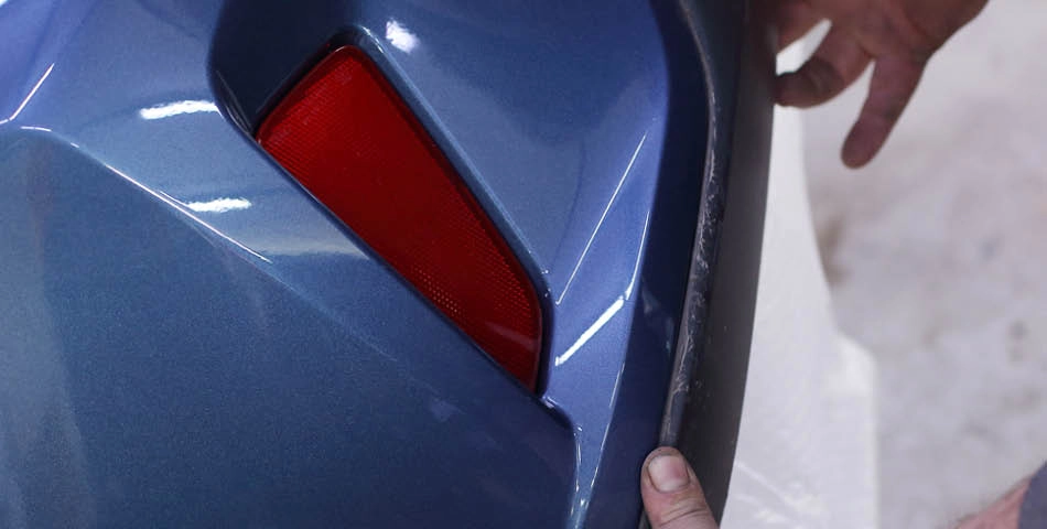 Close-up of a person’s hand touching the edge of a blue car bumper with a red reflector.