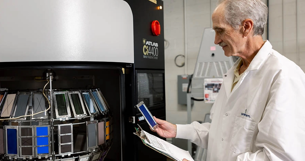 A man in a lab coat examines a smartphone panel next to a large testing machine with multiple devices at a laboratory facility.