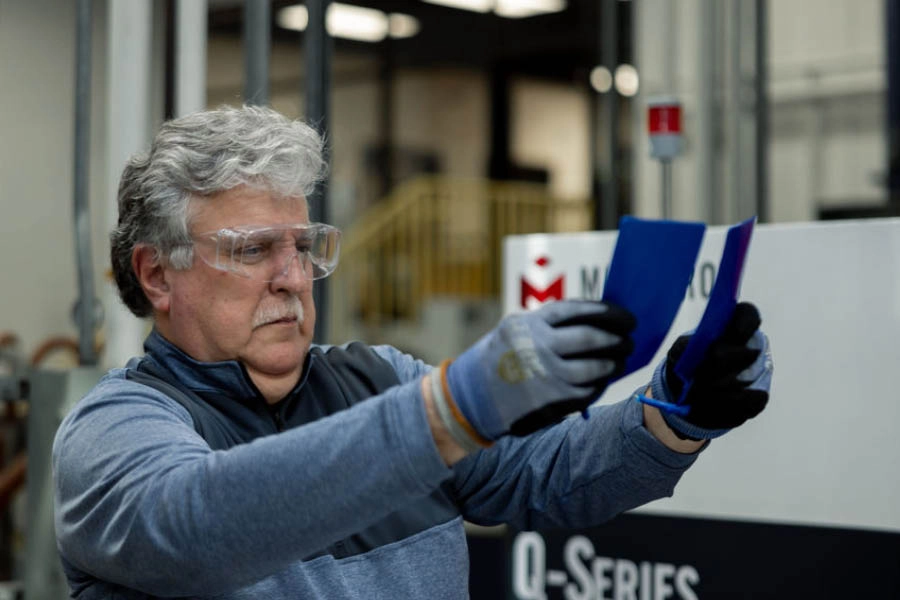 A man wearing safety glasses and gloves inspects two colored sheets in an industrial setting near machinery labeled "Q-Series.