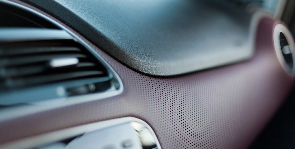Close-up view of a car dashboard showing an air vent, part of the instrument panel, and perforated textured material.