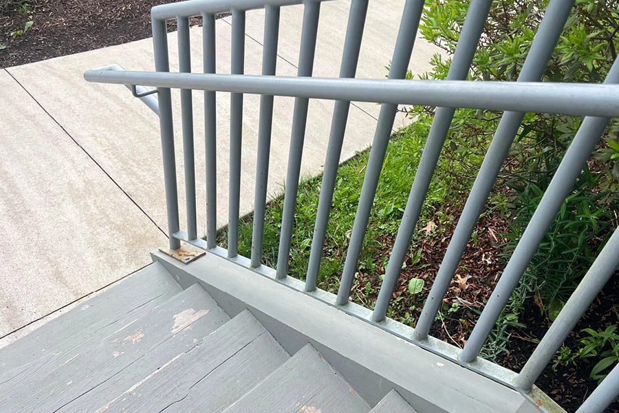 Gray metal handrail and vertical bars beside outdoor steps, with concrete walkway and green bushes visible below.