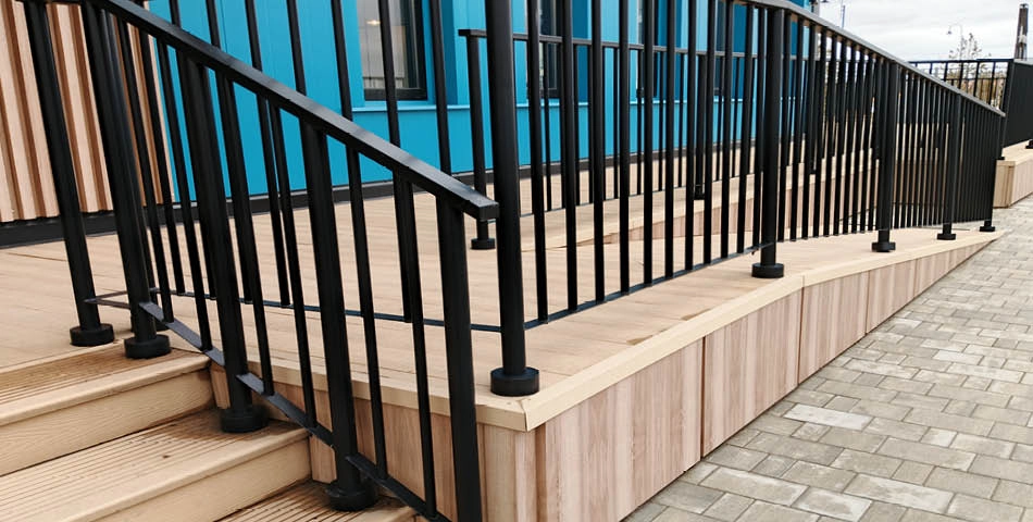 A wooden wheelchair ramp with black metal handrails, next to a building with blue walls and windows, beside a paved walkway.