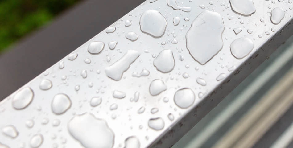 Close-up of water droplets scattered on a smooth, metallic surface, likely after recent rainfall.