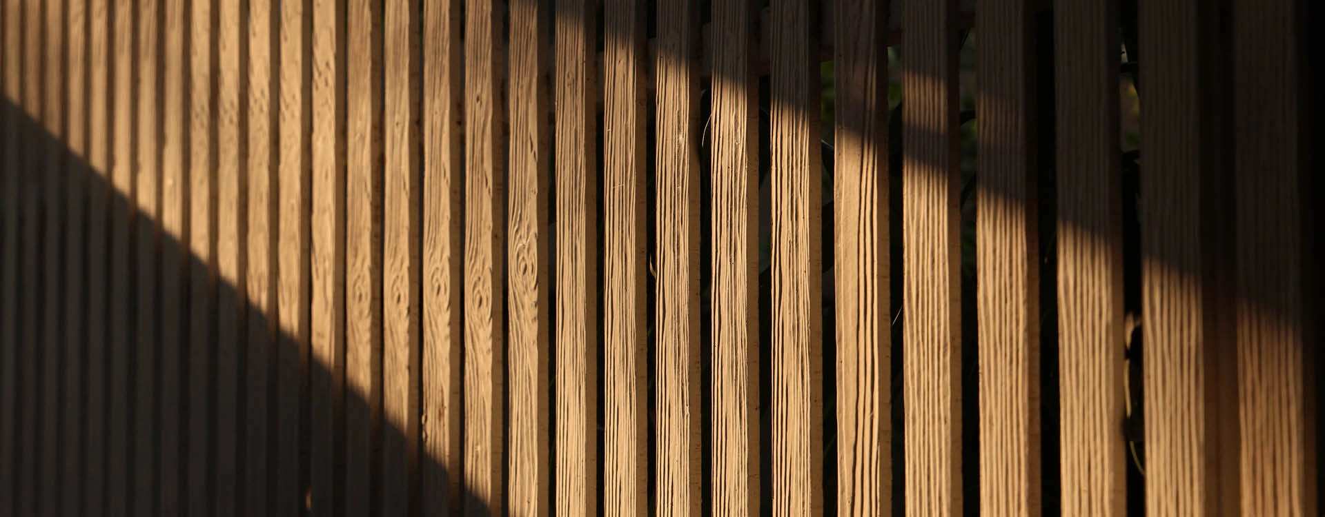 Vertical wooden slats are partially illuminated by sunlight, creating a clear diagonal shadow across the fence.