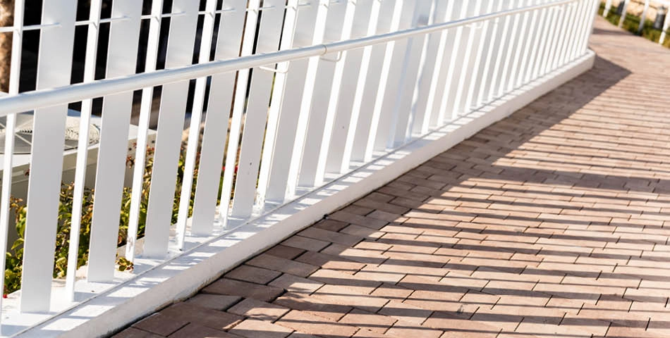 A white metal railing casts striped shadows on a walkway made of rectangular tan bricks in sunlight.