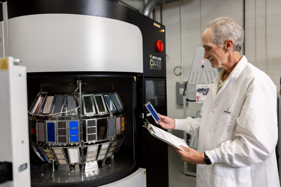 A man in a lab coat examines color samples and records data beside a large industrial color testing machine in a laboratory setting.